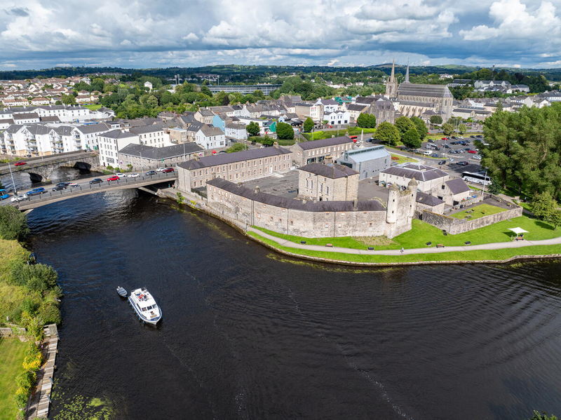 Aerial view of a historic stone castle beside a river, with a boat approaching a bridge and a town with churches and houses in the background.