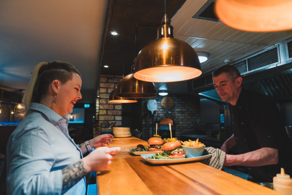 chef handing over two plates of burgers and chips to smiling waitress.